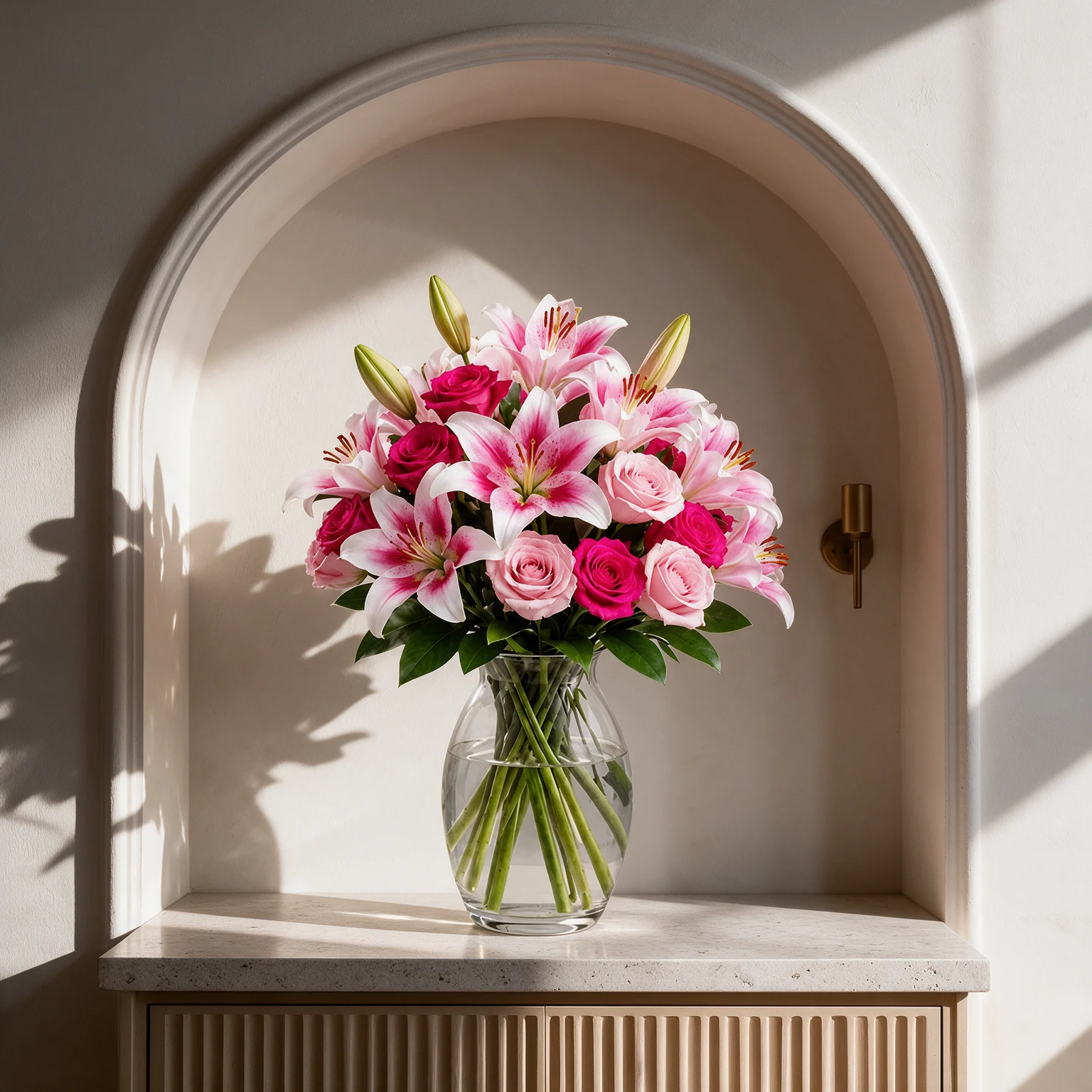 Fuchsia roses, blush pink roses and pink lilies with green foliage arranged in a clear glass vase.