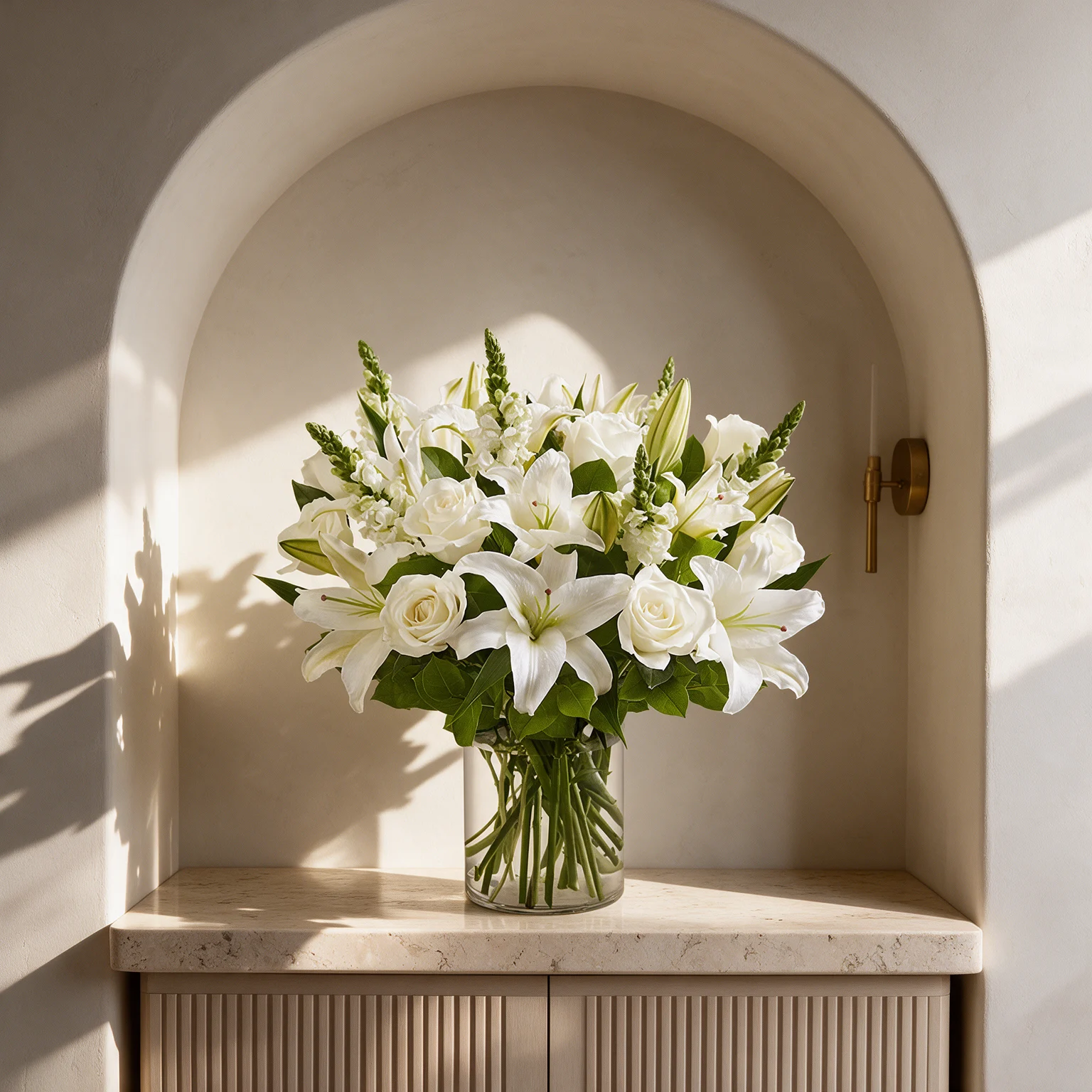 White lilies and ivory roses arranged with delicate white filler flowers and lush green foliage in a clear glass vase.