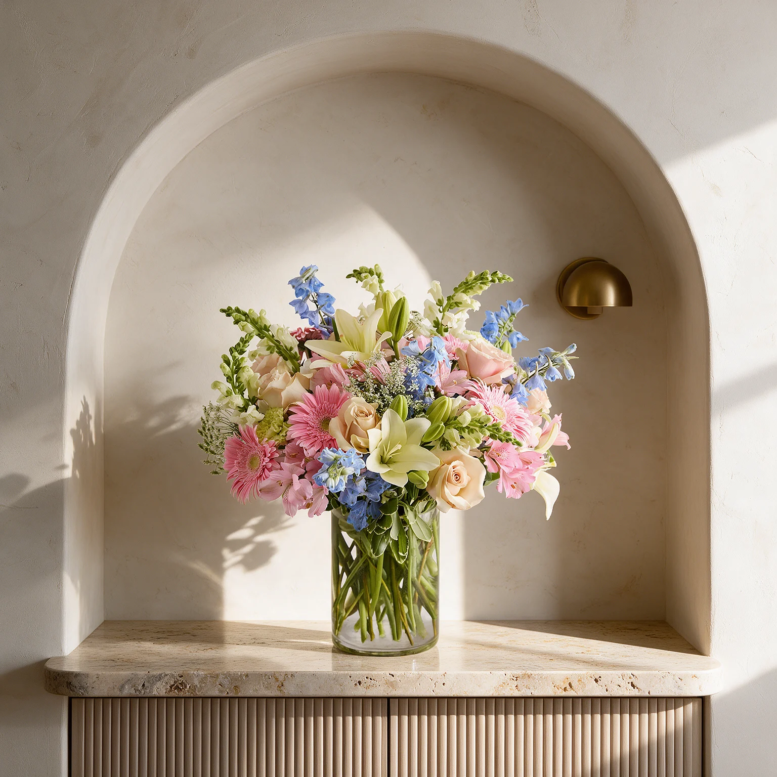 Pink roses and gerberas, white lilies and matthiola, and blue delphiniums arranged with fresh green foliage in a clear glass vase.