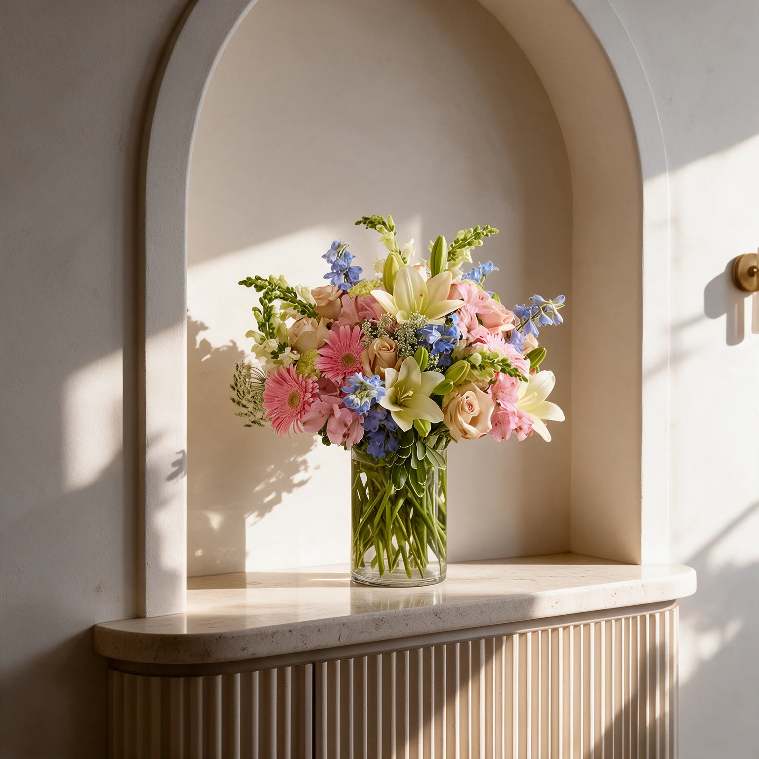 Pink roses and gerberas, white lilies and matthiola, and blue delphiniums arranged with fresh green foliage in a clear glass vase.