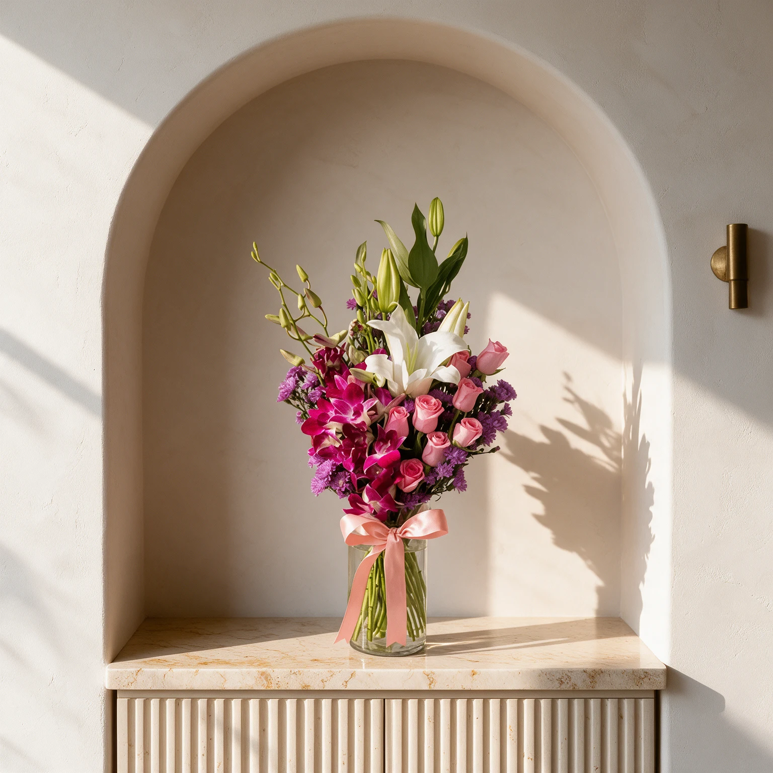 Pink roses, white lilies and magenta orchids with purple filler flowers arranged in a clear glass vase with a pink bow.