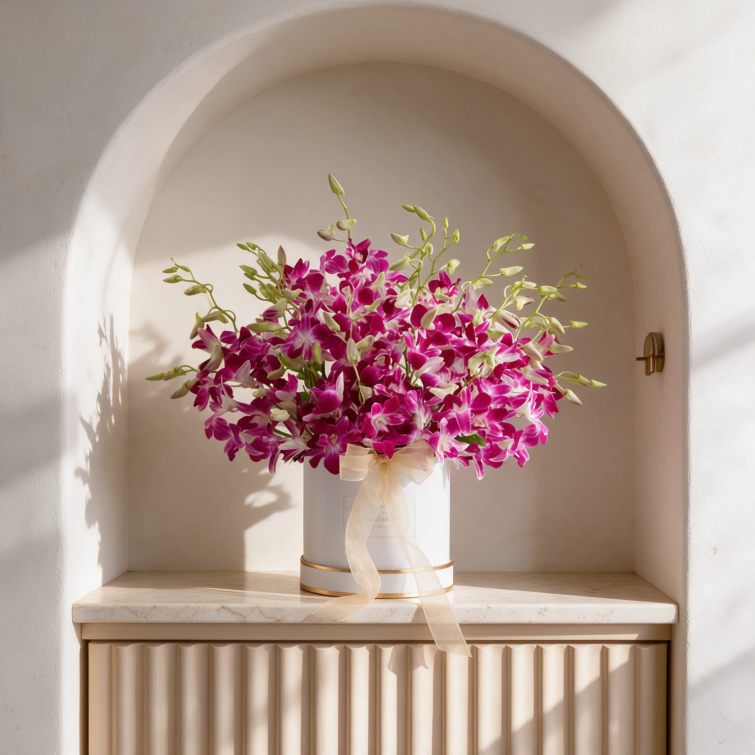 Vibrant magenta orchids with white centres arranged in a white cylindrical hatbox with gold trim and a cream ribbon.